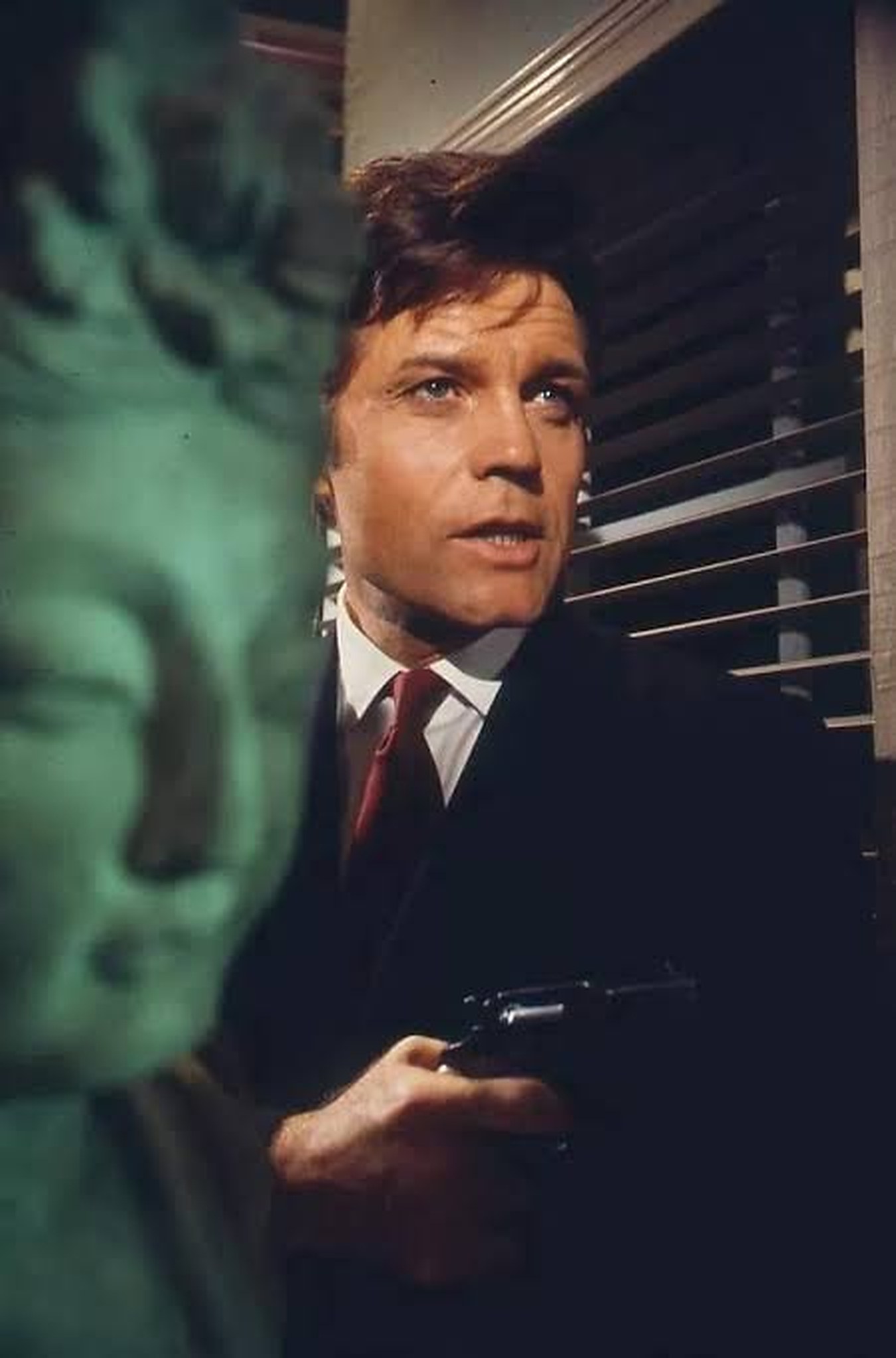Jack Lord, alias Steve McGarrett, revolver à la main, derrière une sculpture verte — Hawaï Police d'État